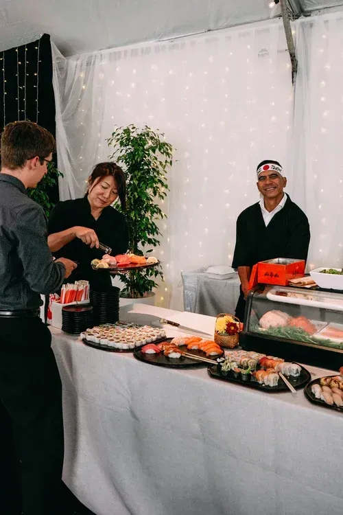 People serving food at a buffet table with trays of appetizers in a bright event tent
