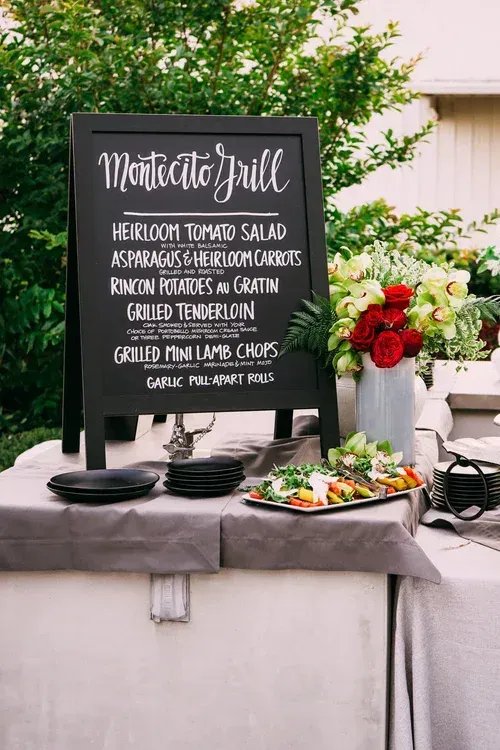 Black wedding menu sign with food table and red floral centerpiece outdoors