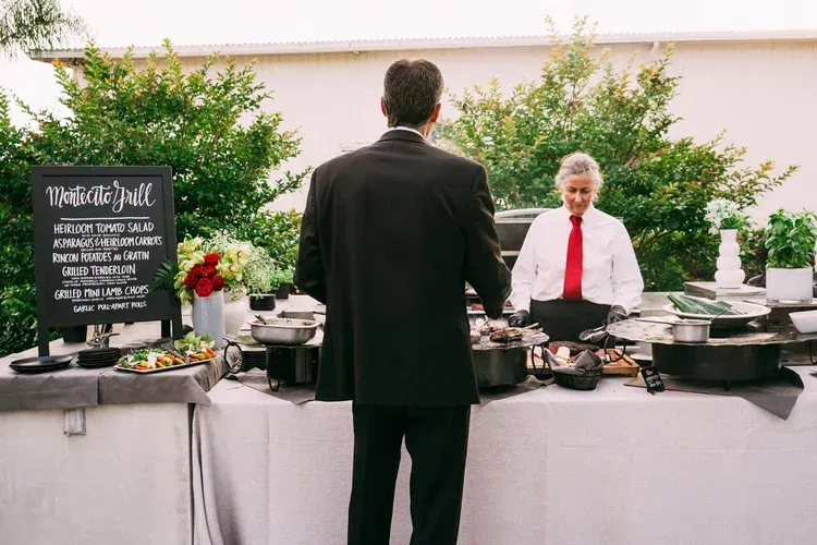 Outdoor buffet with a chef behind serving dishes and a guest in a black suit facing him