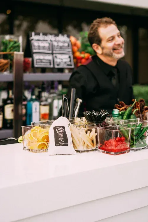 Bartender smiling behind a bar with cocktail ingredients, bottles, and glass bowls of fruit and herbs
