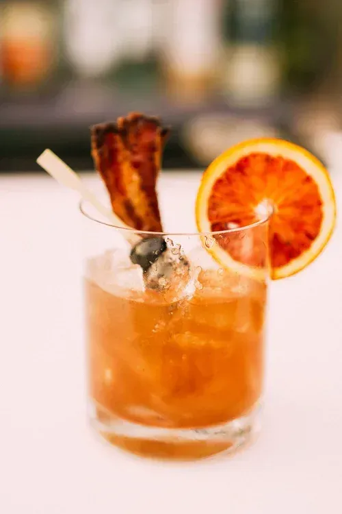 Orange cocktail with a dried citrus slice and bacon garnish in a glass on a bar counter