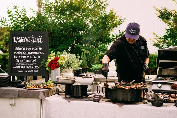 Outdoor chef serving food at a buffet station beside a menu sign and grill setup
