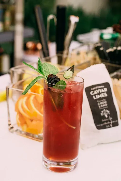 Red cocktail with blackberry and lime garnish beside a napkin on a bar counter