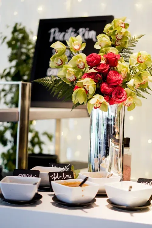 Floral arrangement in a silver vase beside bowls and cups on a table with blurred festive lights.