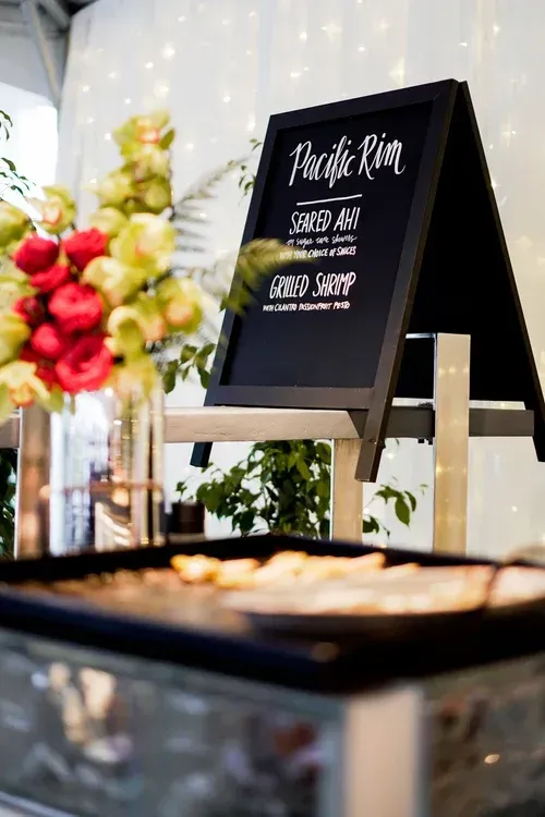 Black chalkboard wedding sign beside a flower arrangement and appetizer tray on a decorated table