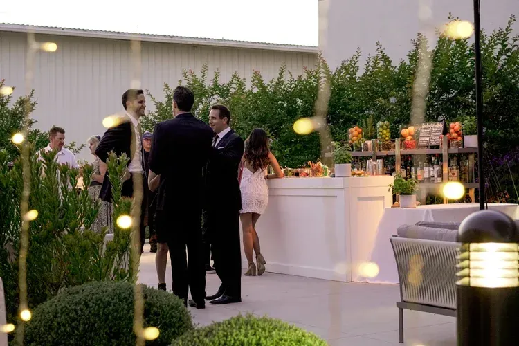 Guests mingle at an outdoor evening reception beside a white bar and lush greenery.