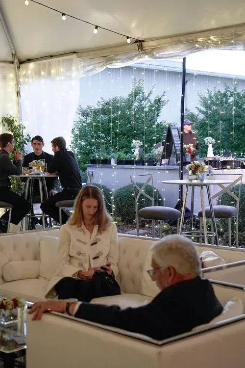 People seated under a clear tent at an outdoor event, with string lights and a serving table in the background.