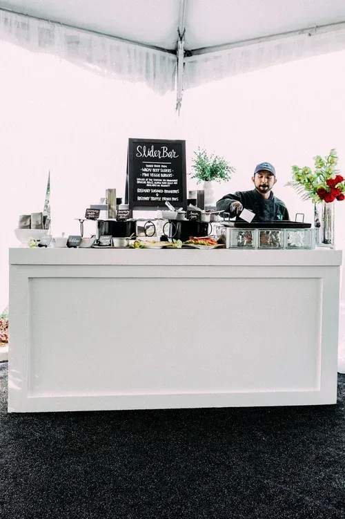 Coffee stand with a barista behind a white counter, chalkboard menu, espresso equipment, and flowers in a bright tent