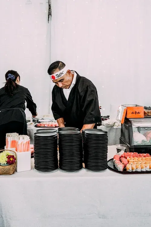 Caterer arranging food trays behind a white table with stacked black plates and orange drink cartons