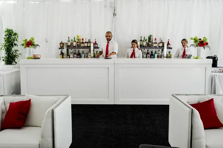 White reception bar with two staff behind it, floral arrangements, and red cushions in a bright event space