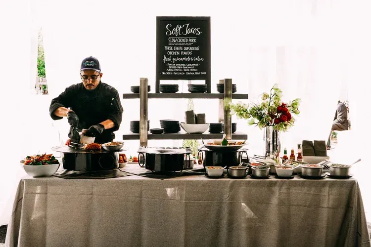 Chef serving food at a buffet table with pots, flowers, and a menu sign