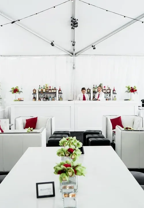 White banquet tent with white tables, black chairs, red accents, and a bar setup in the background