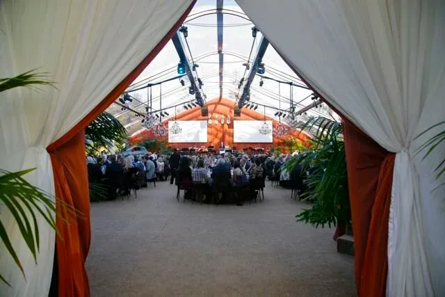 Crowd seated in a tented event space, seen through white and orange drapes.