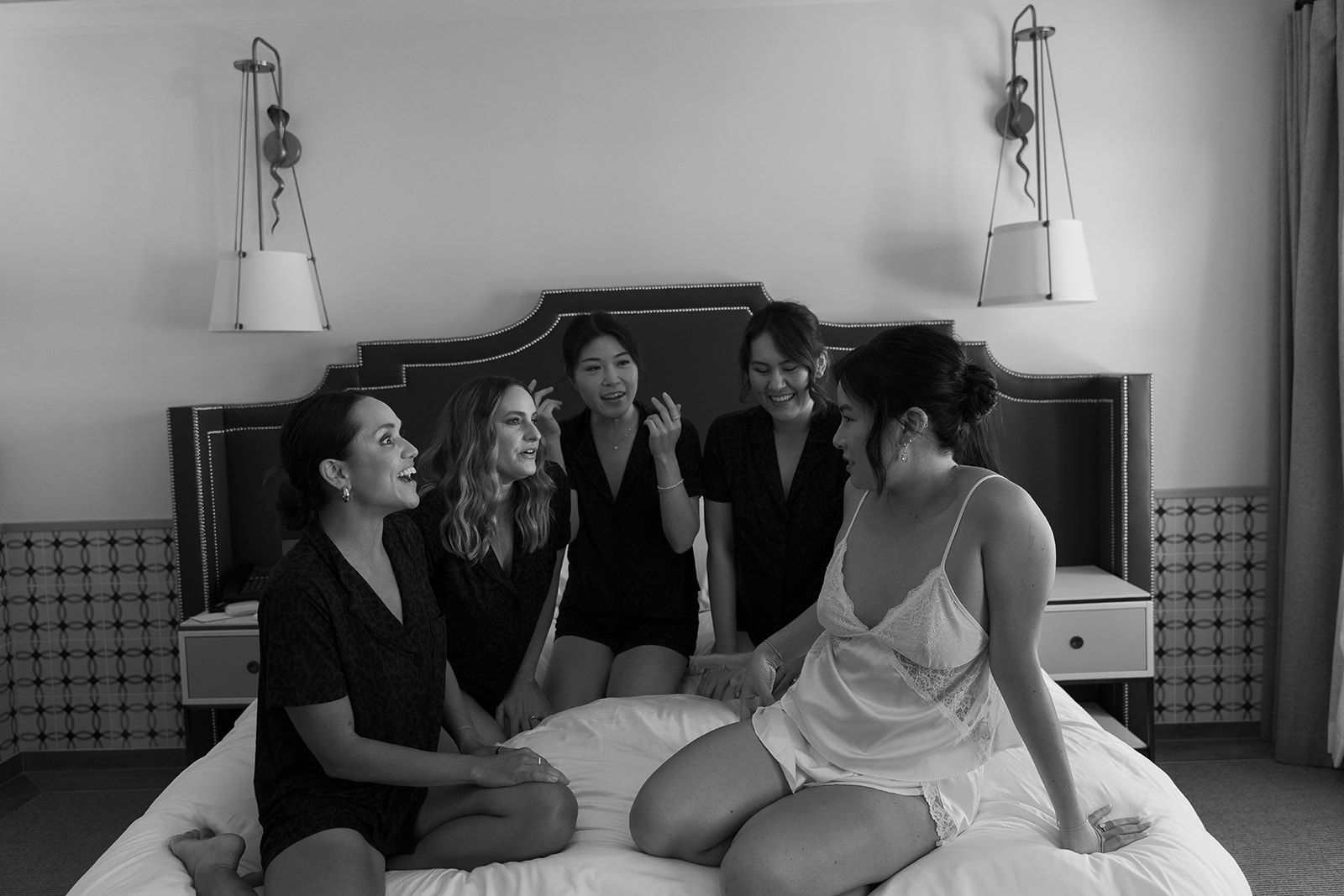 Four women sitting on a bed in a bedroom, chatting and smiling.