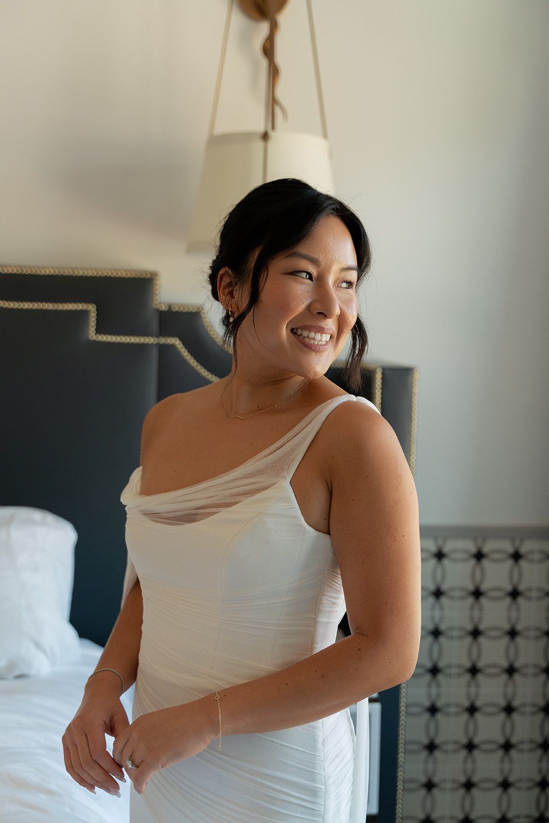 Woman in a white dress smiling by a bed in a softly lit bedroom