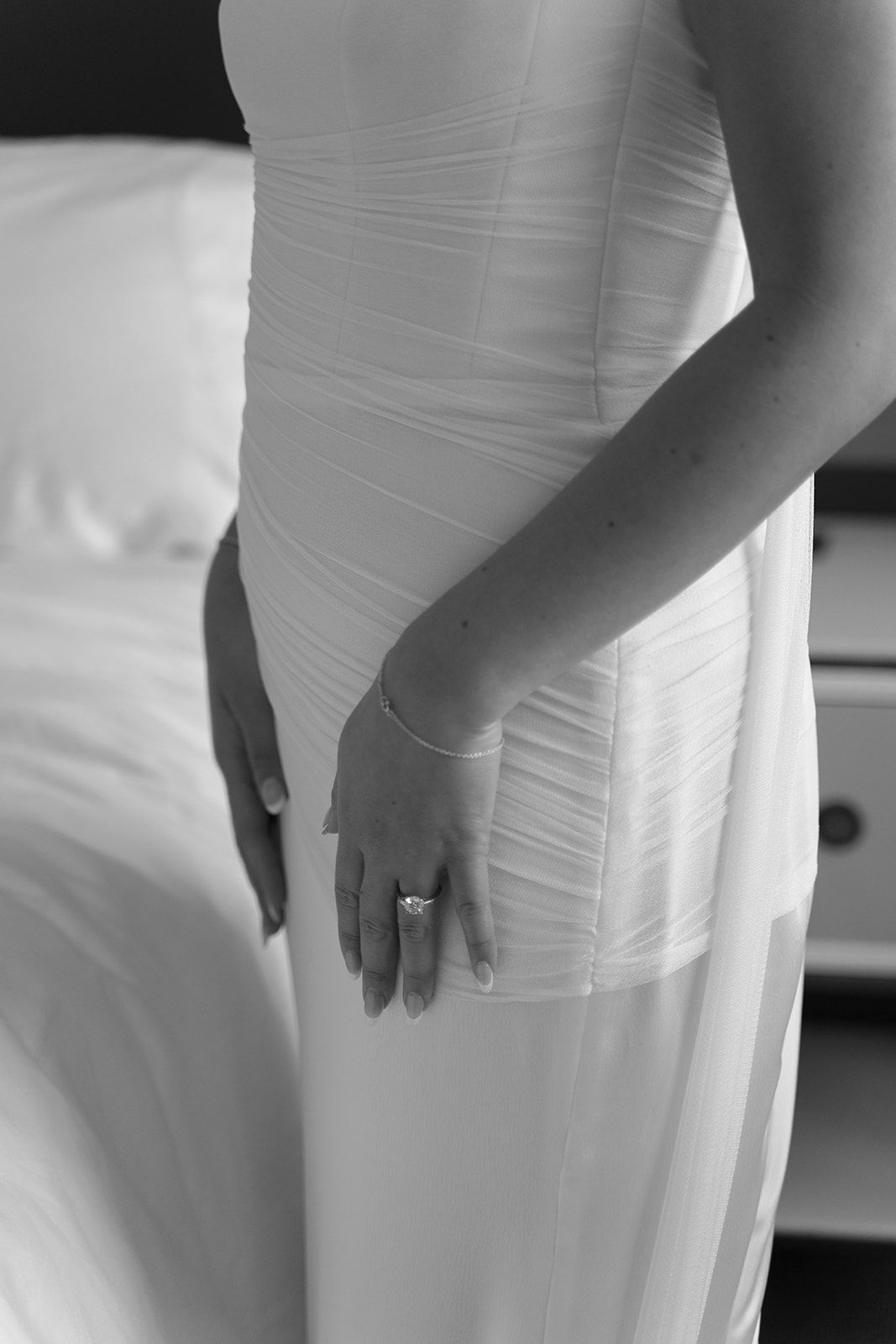 Person in a white dress standing beside a bed, hands resting at their side.