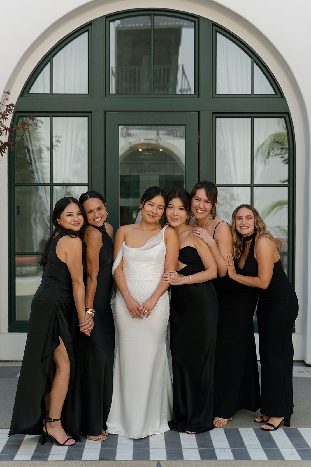 Six people posing in formal black and white outfits in front of a large arched window.