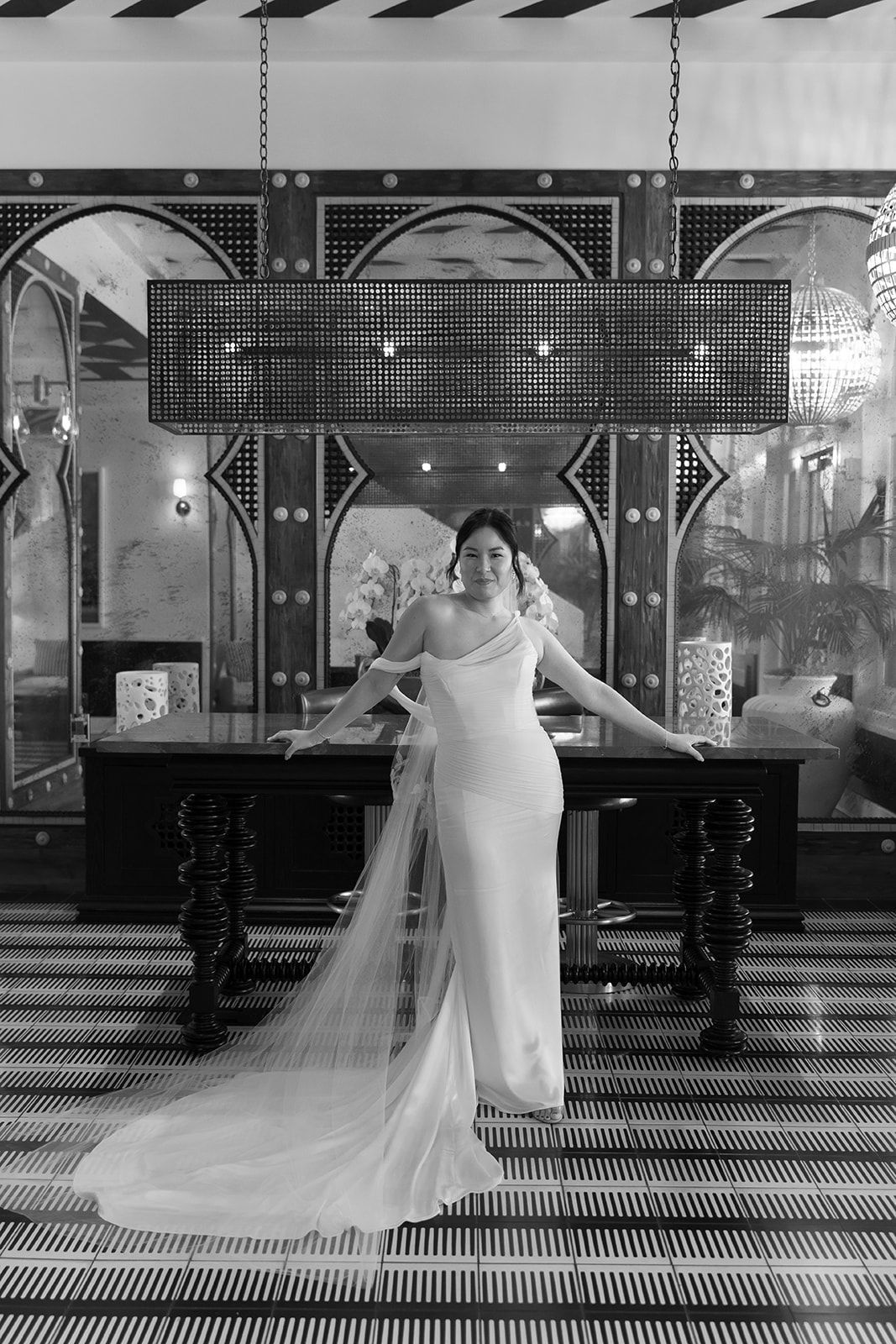 Woman in a white gown standing on a patterned floor in an ornate hall, with a long train behind her.