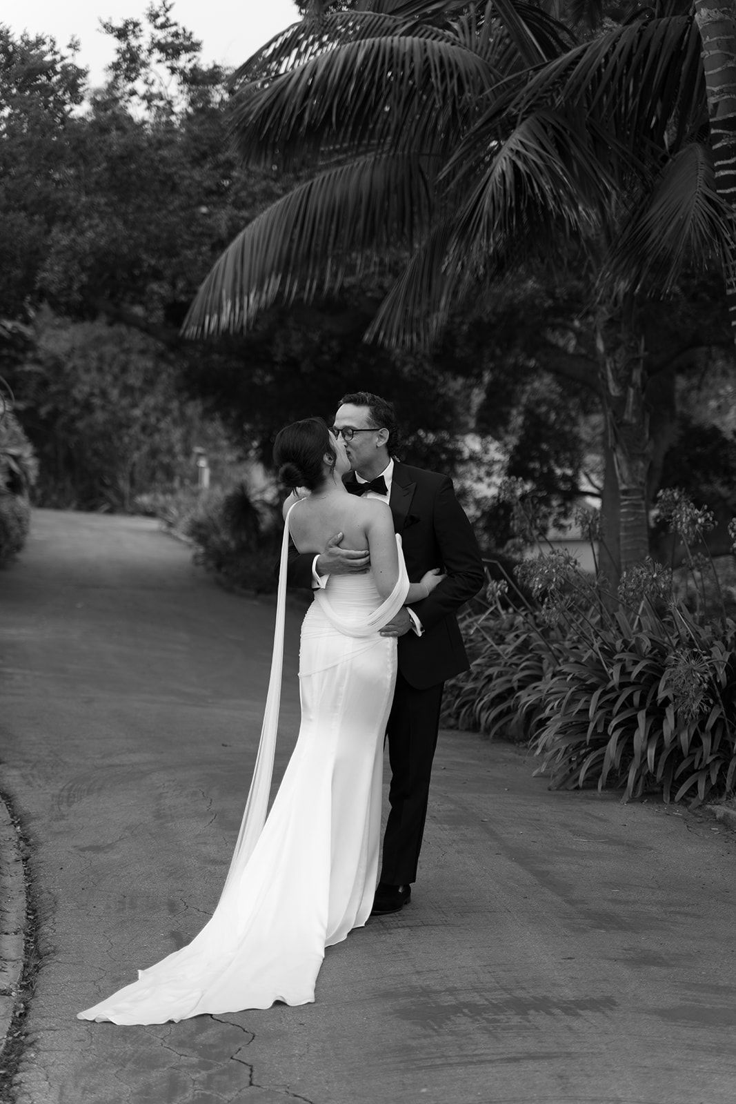 Bride and groom standing together outdoors in a garden path, the bride in a white gown and the groom in a dark suit