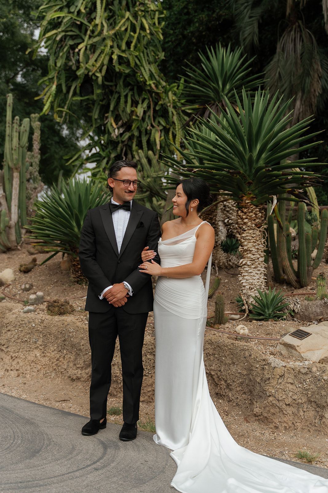 Bride and groom posing in formal attire in a lush garden, smiling at each other.
