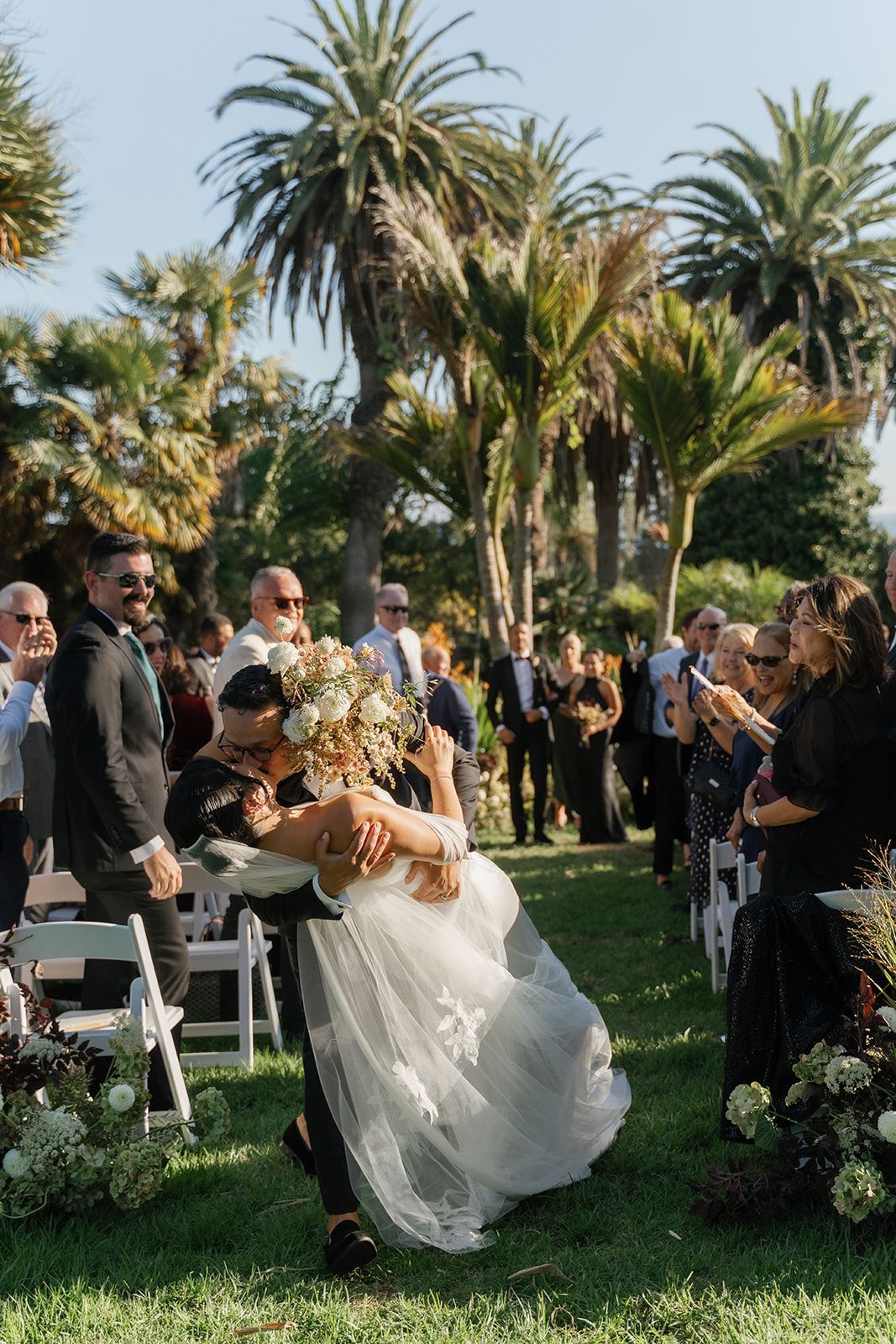 Bride in a white gown being dipped during a wedding ceremony outdoors among guests and palm trees
