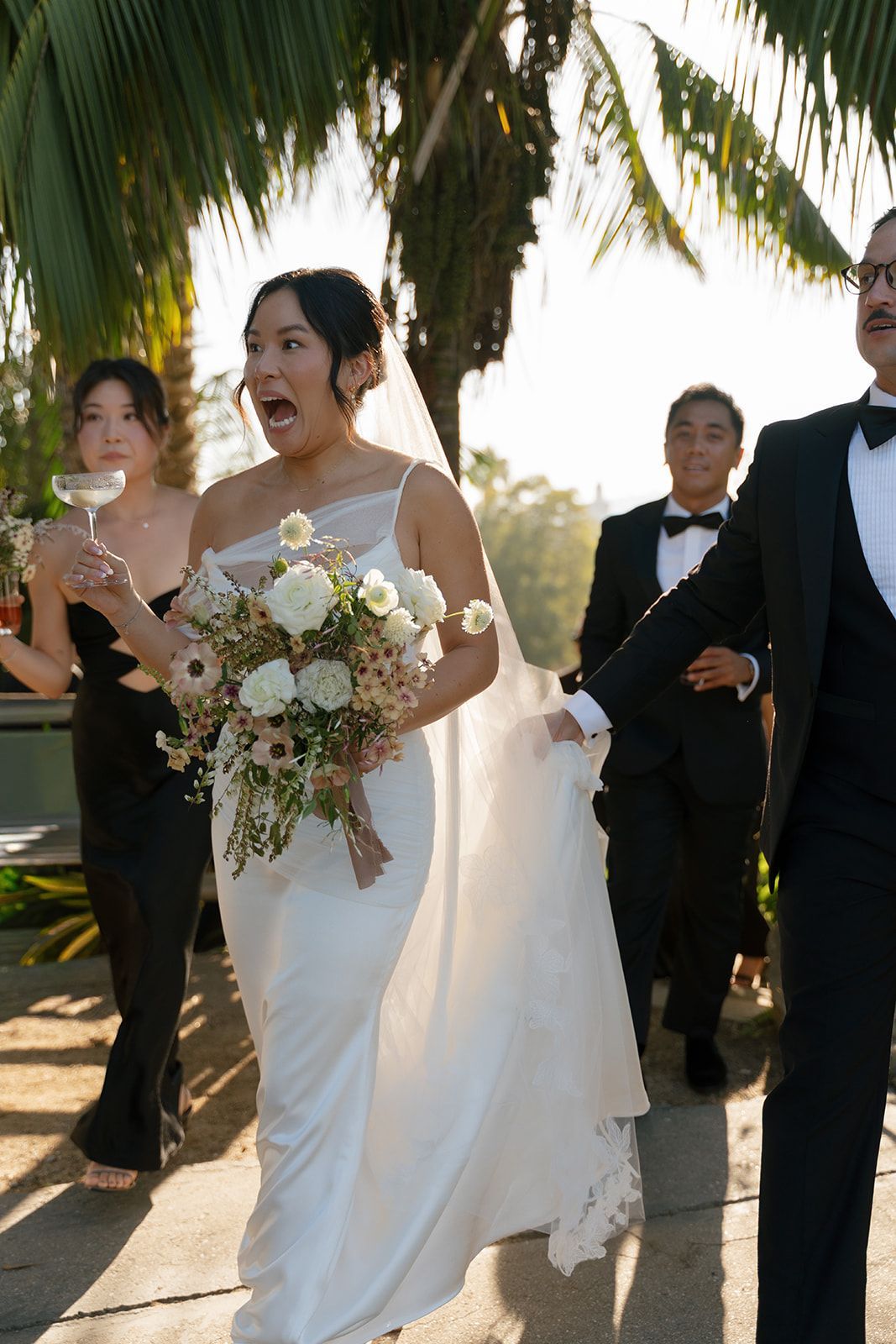 Bride in white gown walking outdoors with bouquet, flanked by attendants in tuxedos under palm trees.