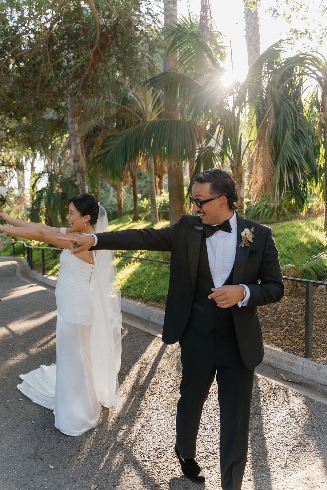 Couple in formal attire posing outdoors on a sunlit path with palm trees, arms extended toward each other.