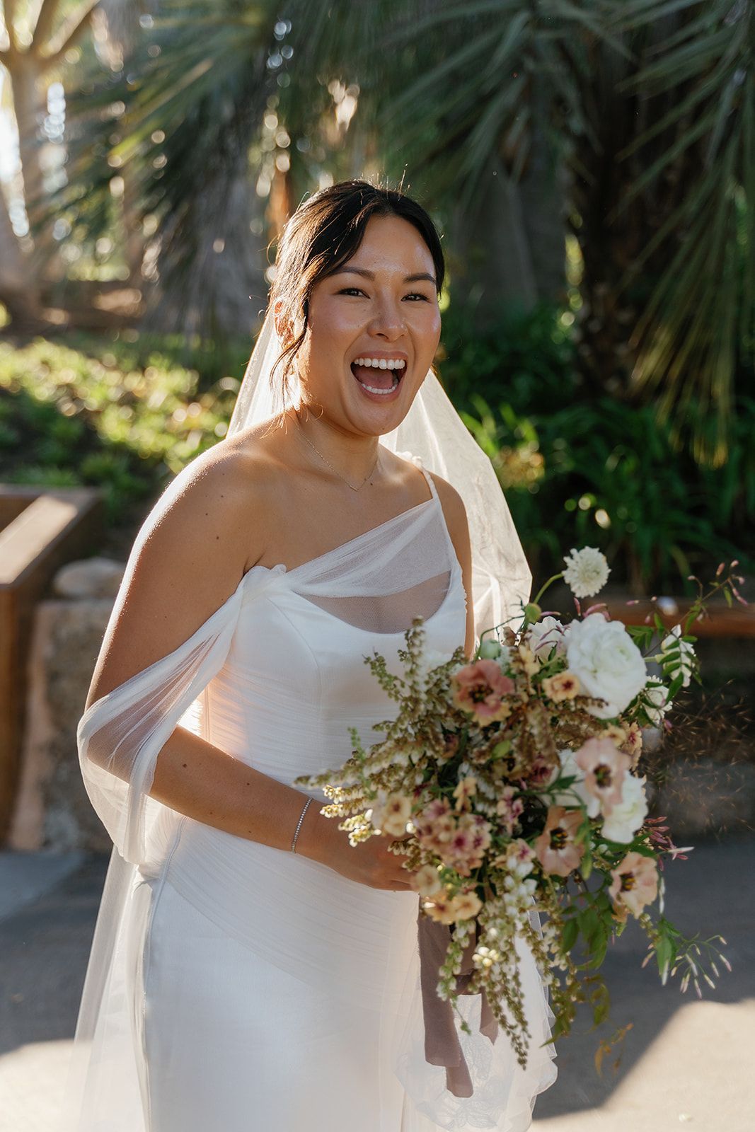 Smiling bride in a white off-shoulder gown holding a bouquet outdoors