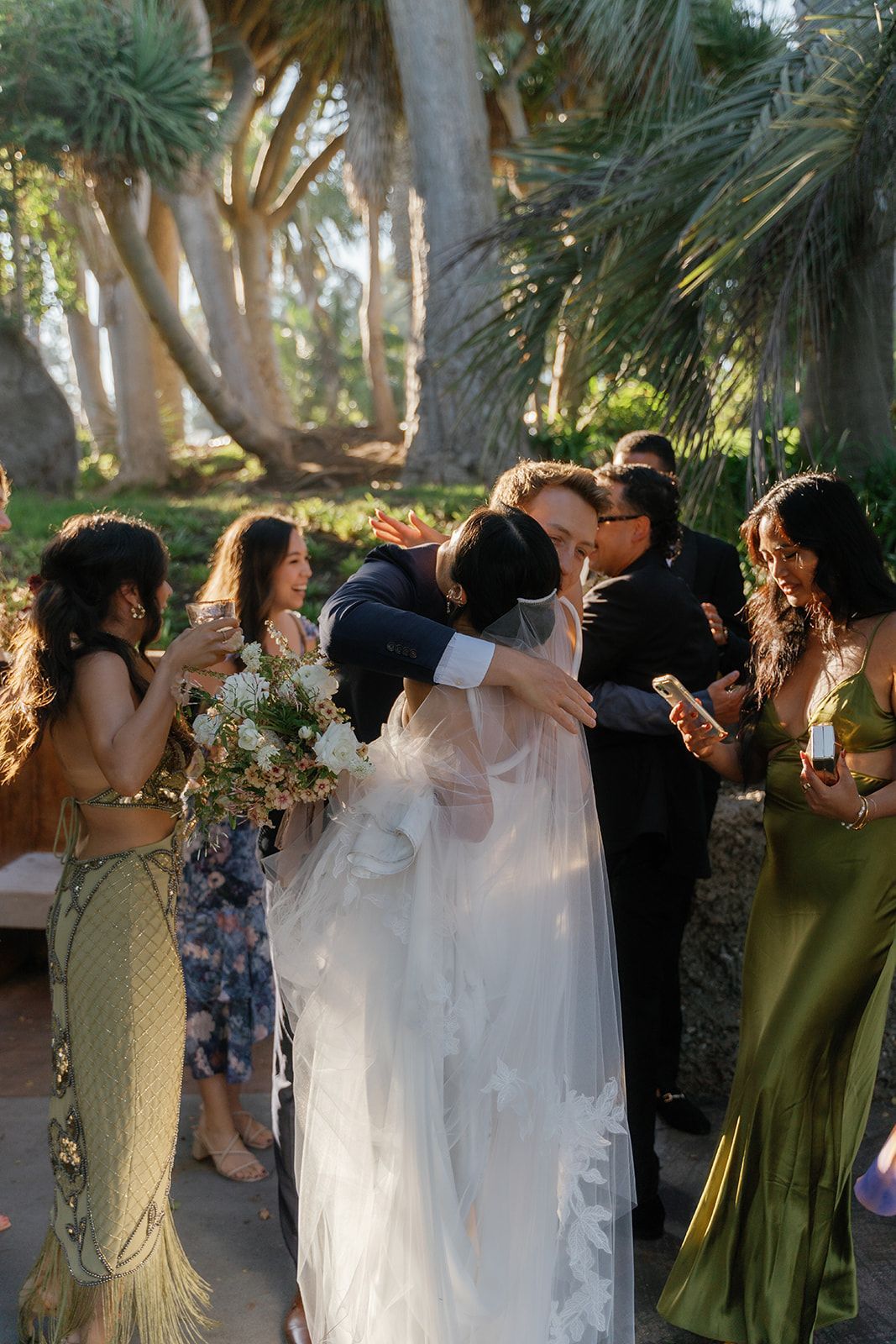 Wedding party embracing outdoors among palm trees, with a bride in a white dress and guests holding flowers.