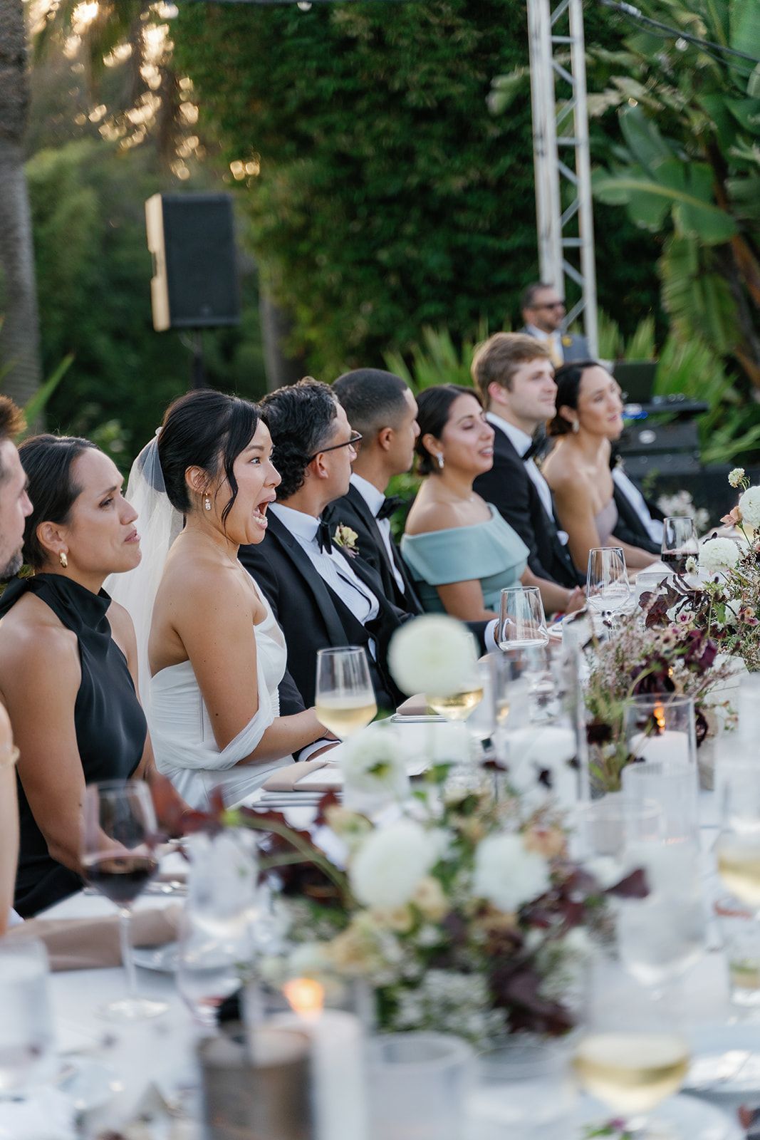 Guests seated at a long outdoor dinner table in a garden, dressed in formal attire.