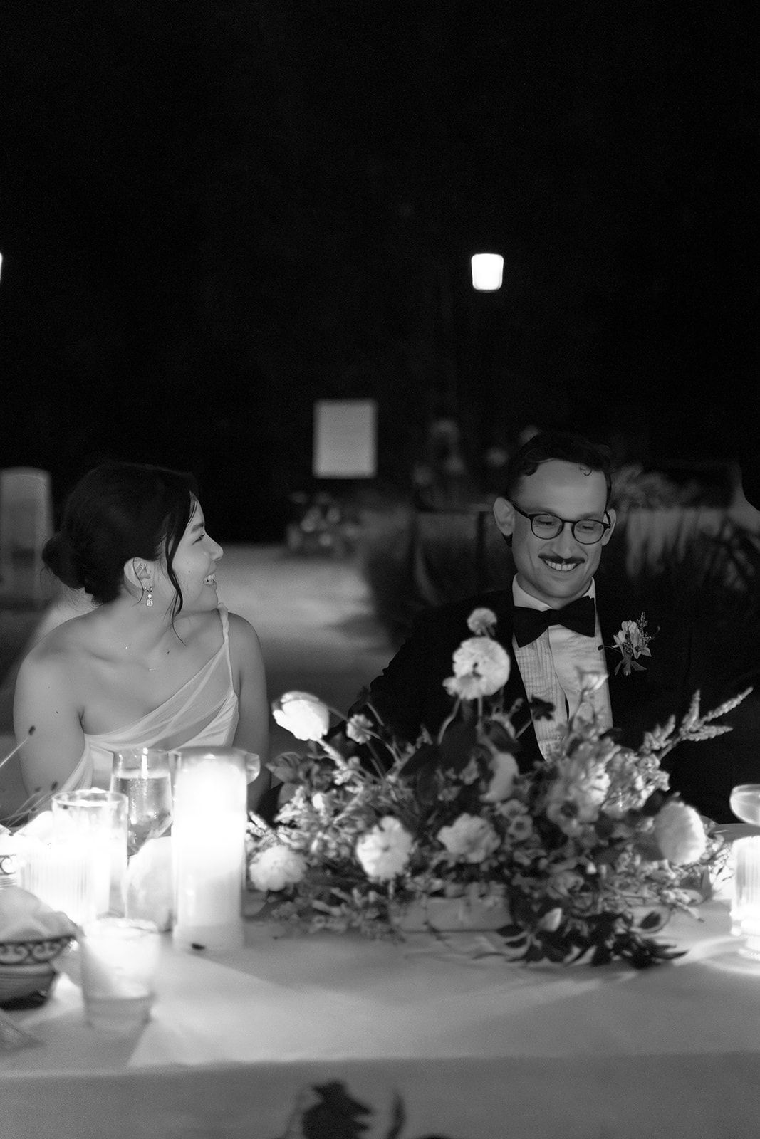 Dinner guests seated at a candlelit outdoor table with flowers and drinks at night