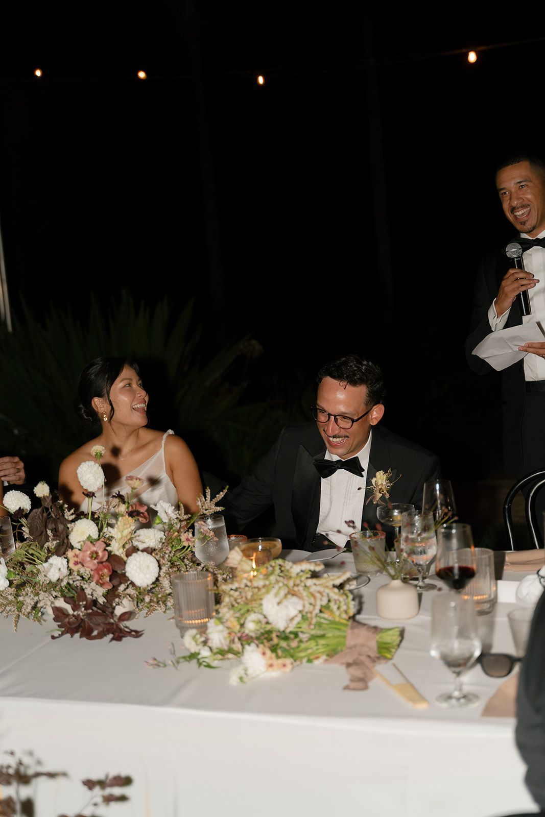 Formal dinner toast with seated couple at a decorated table and a man speaking beside them.