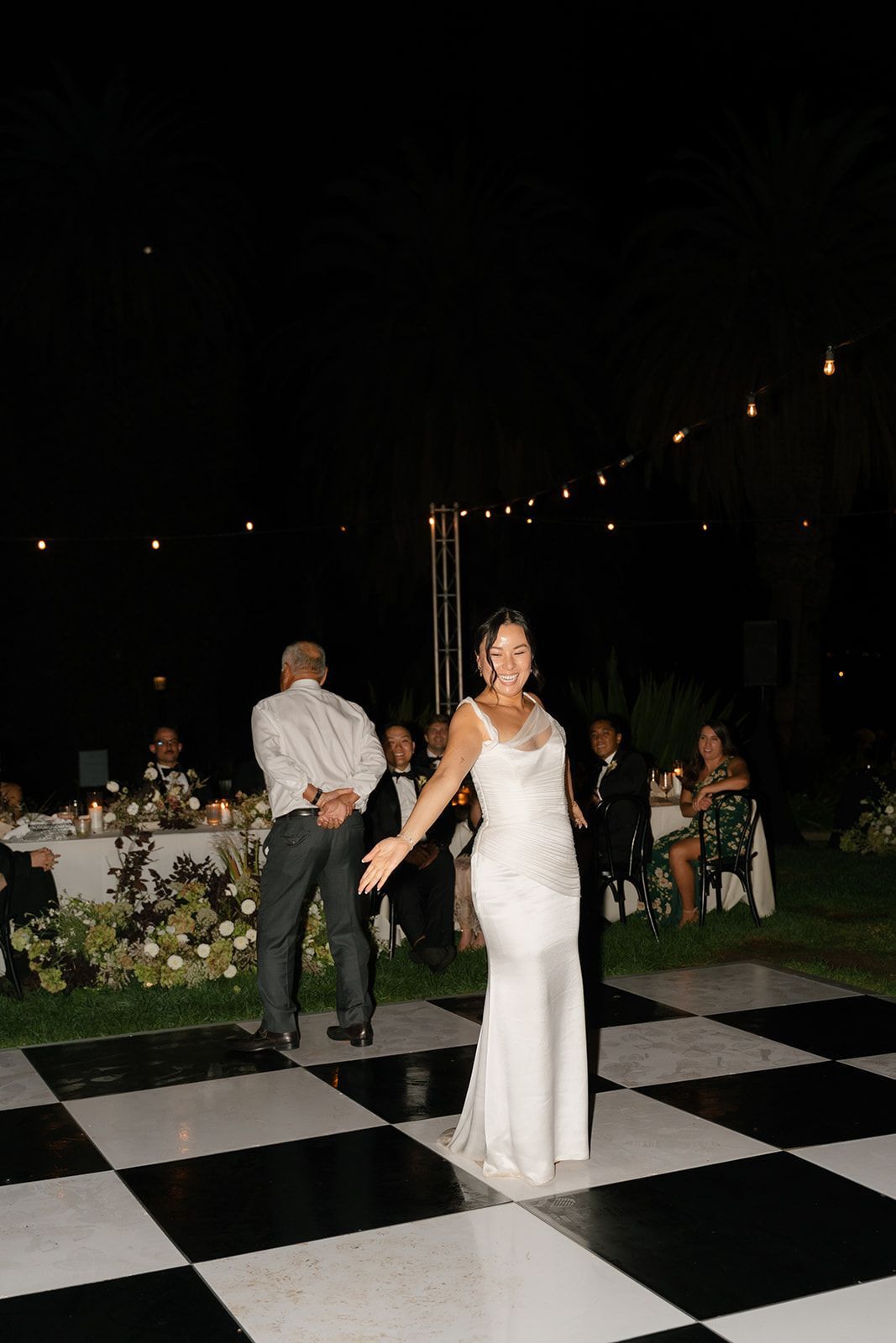 Bride in white dress stands on a checkered dance floor at an outdoor evening wedding reception.