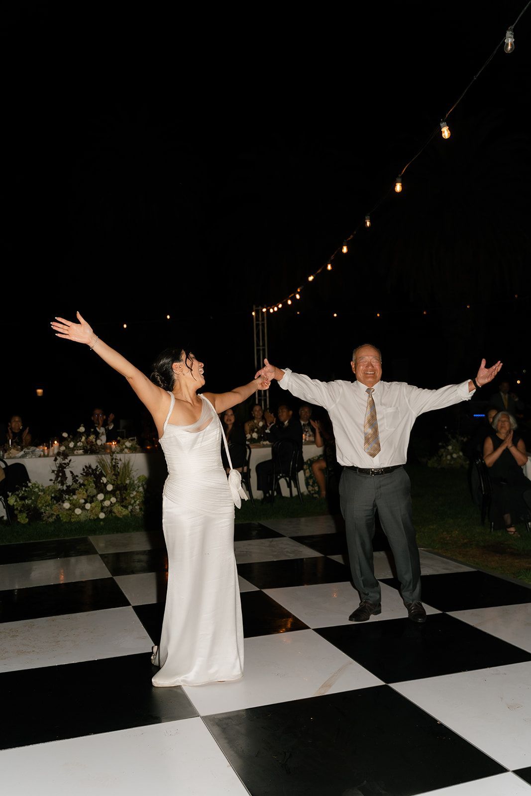 Couple celebrating on a black-and-white dance floor at an outdoor evening reception, arms raised
