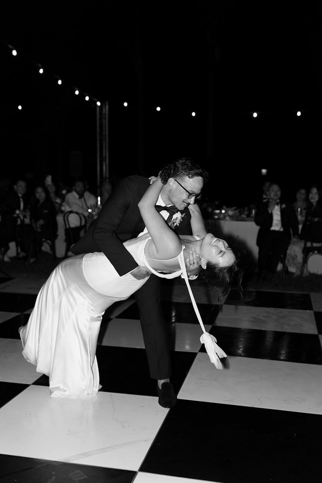 Couple dancing on a black-and-white checkered floor under string lights at an outdoor event