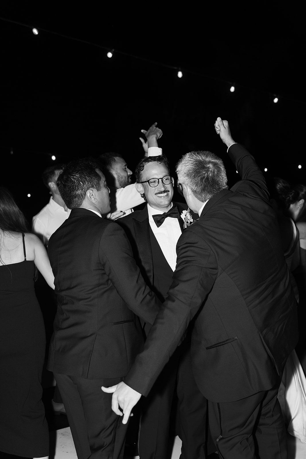 Three men in tuxedos celebrate on a dance floor, raising their hands under string lights.