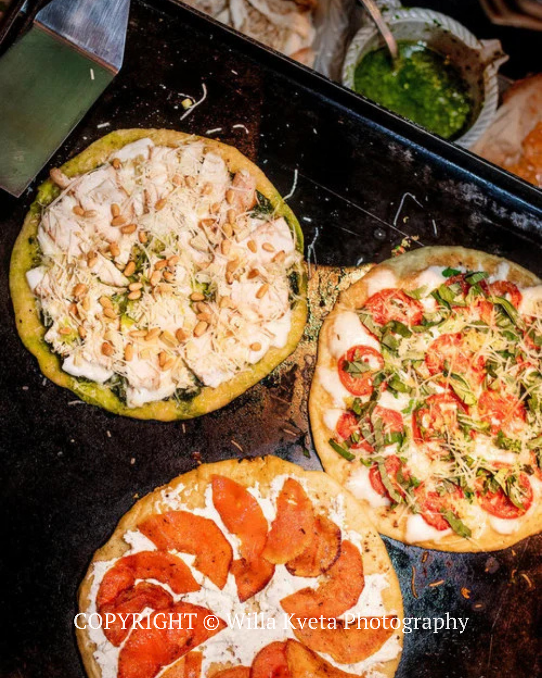 Three topped pizza/flatbread rounds cooking on a dark griddle, viewed from above