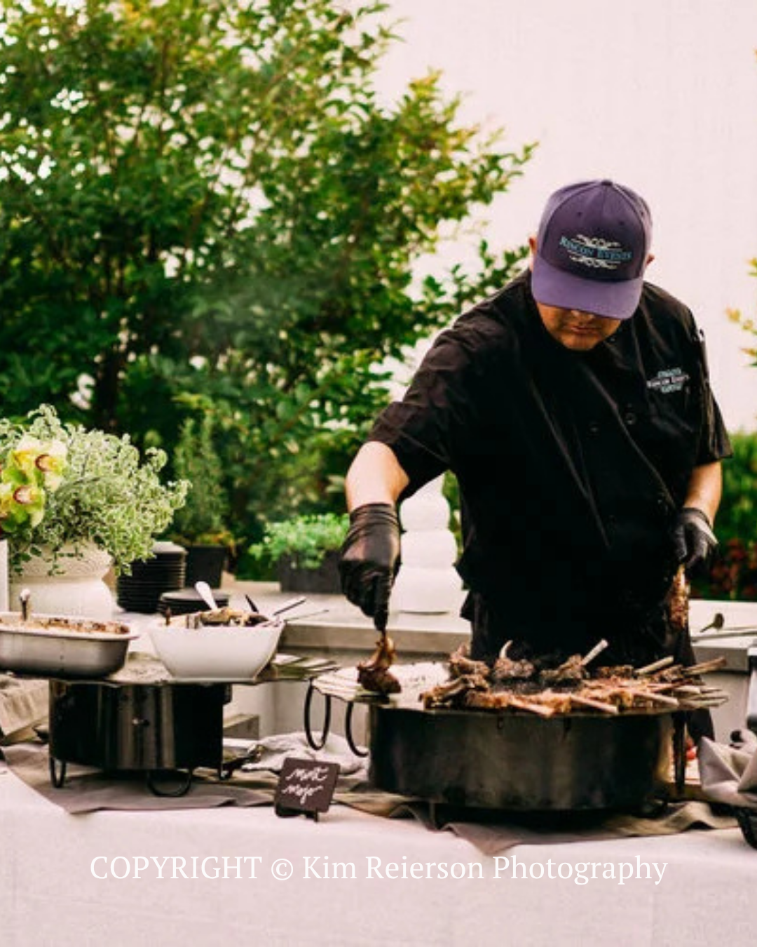 Chef grilling meat outdoors at a barbecue station, wearing a black shirt and cap, with trees in the background