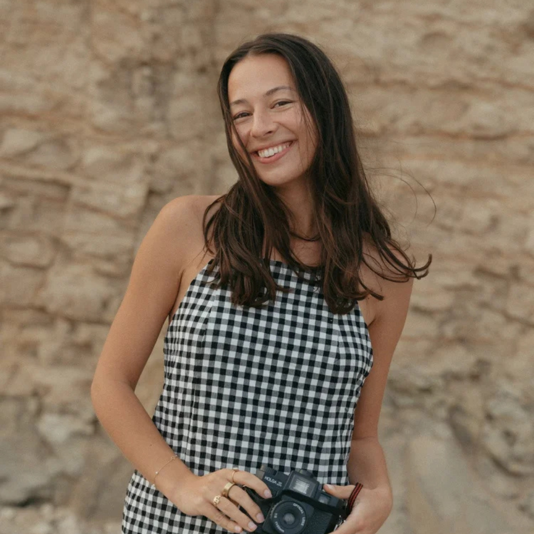 Woman smiling in a black-and-white checkered top, holding a camera against a rocky backdrop