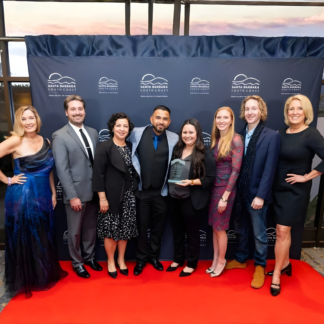 Group of people posing on a red carpet in front of a blue event backdrop, holding an award.