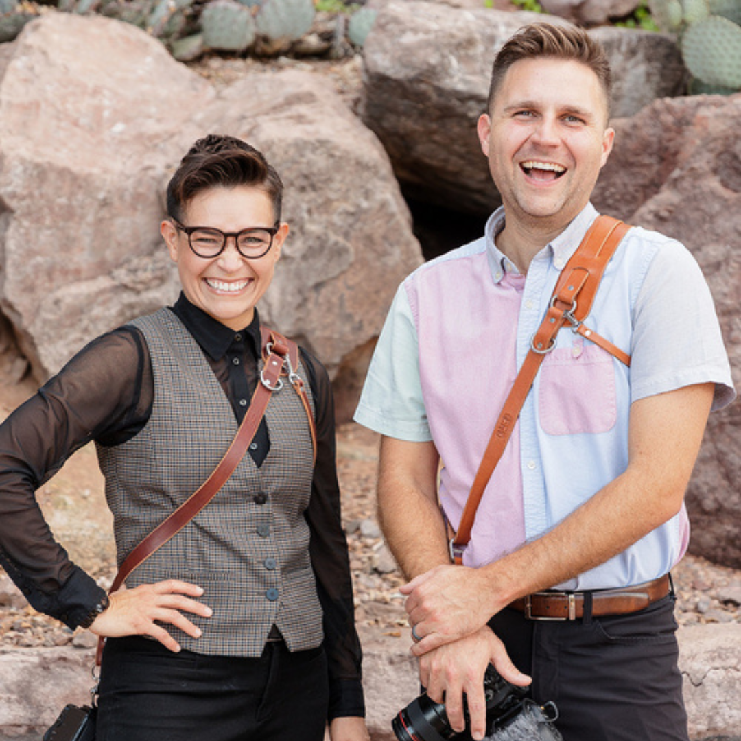 Two smiling people posing outdoors in front of large rocks, wearing casual shirts and crossbody straps.