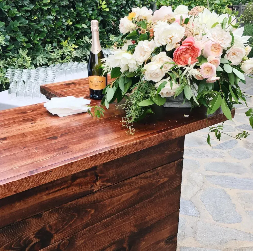 Wooden table with champagne, folded napkins, and a lush peach-and-white floral arrangement outdoors.