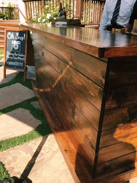 Rustic wooden bar counter outdoors with a chalkboard sign beside it and a person standing behind the counter.