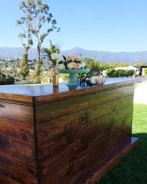 Wooden outdoor bar table with potted plants on a sunny lawn, mountains in the background