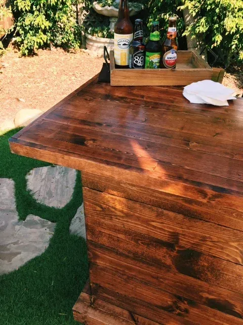 Wooden outdoor table with bottles and a tray on top, beside a patch of artificial grass