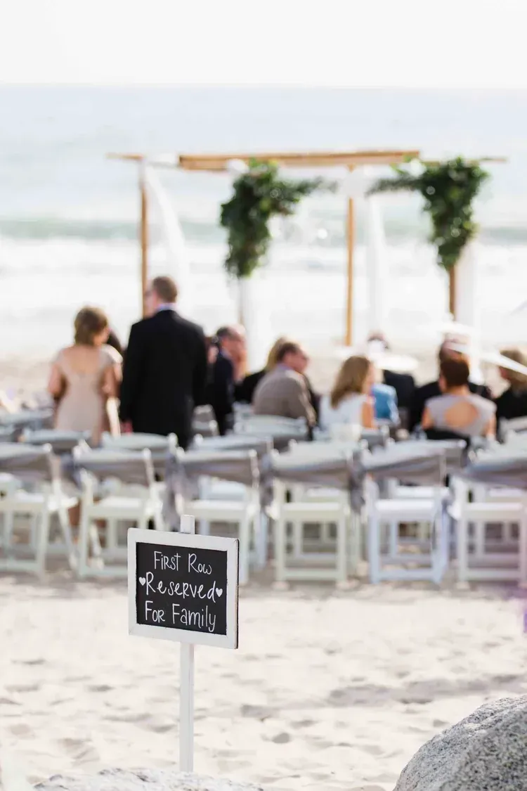 Beach wedding reception with white chairs, floral arch, and guests seated near the water