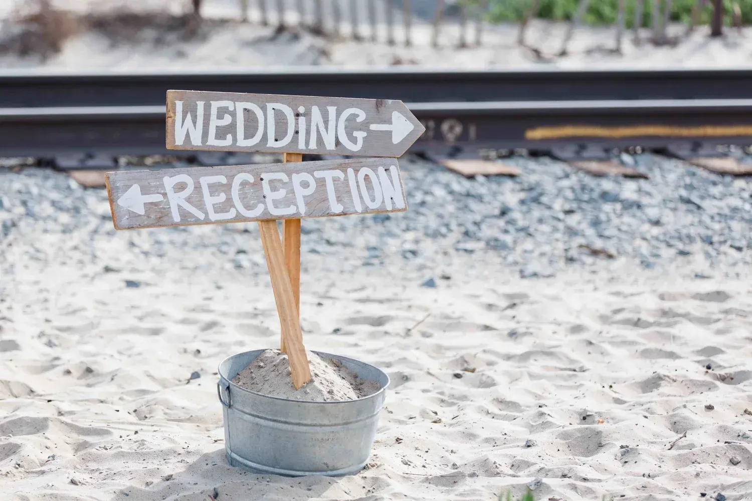 Rustic wedding reception sign in a metal bucket on a sandy beach, with an arrow pointing right.