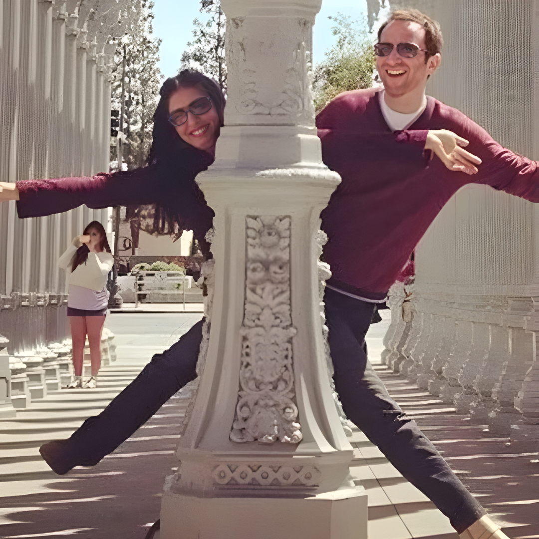 Two people posing playfully around a decorative white column in a sunlit colonnade