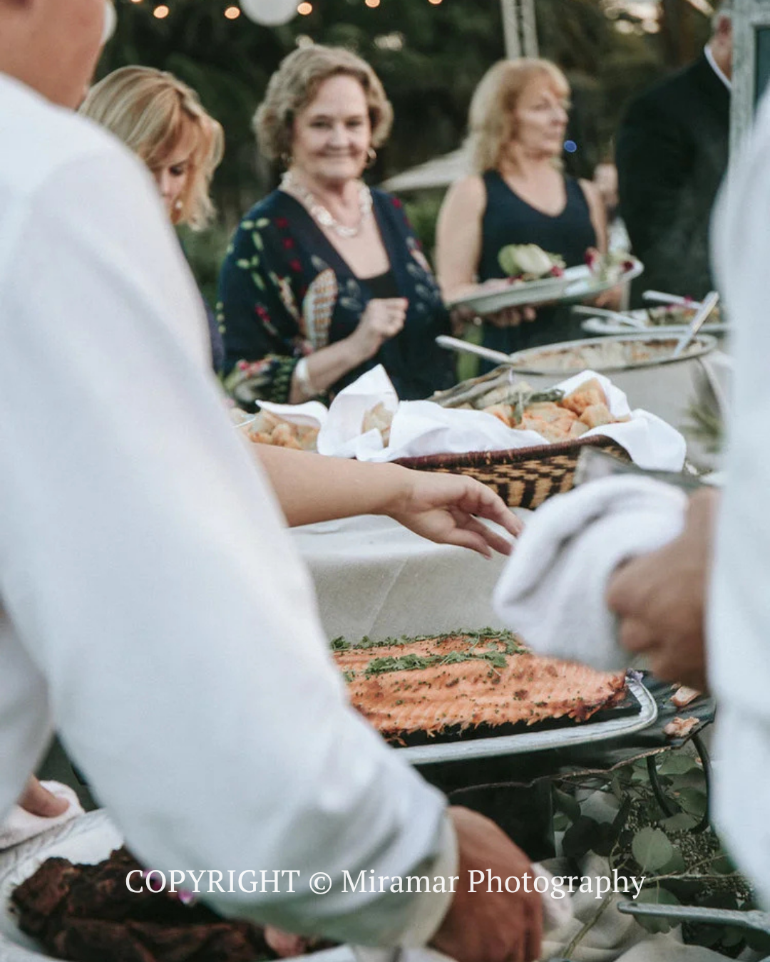 People serving food buffet-style at an outdoor gathering, with guests in the background