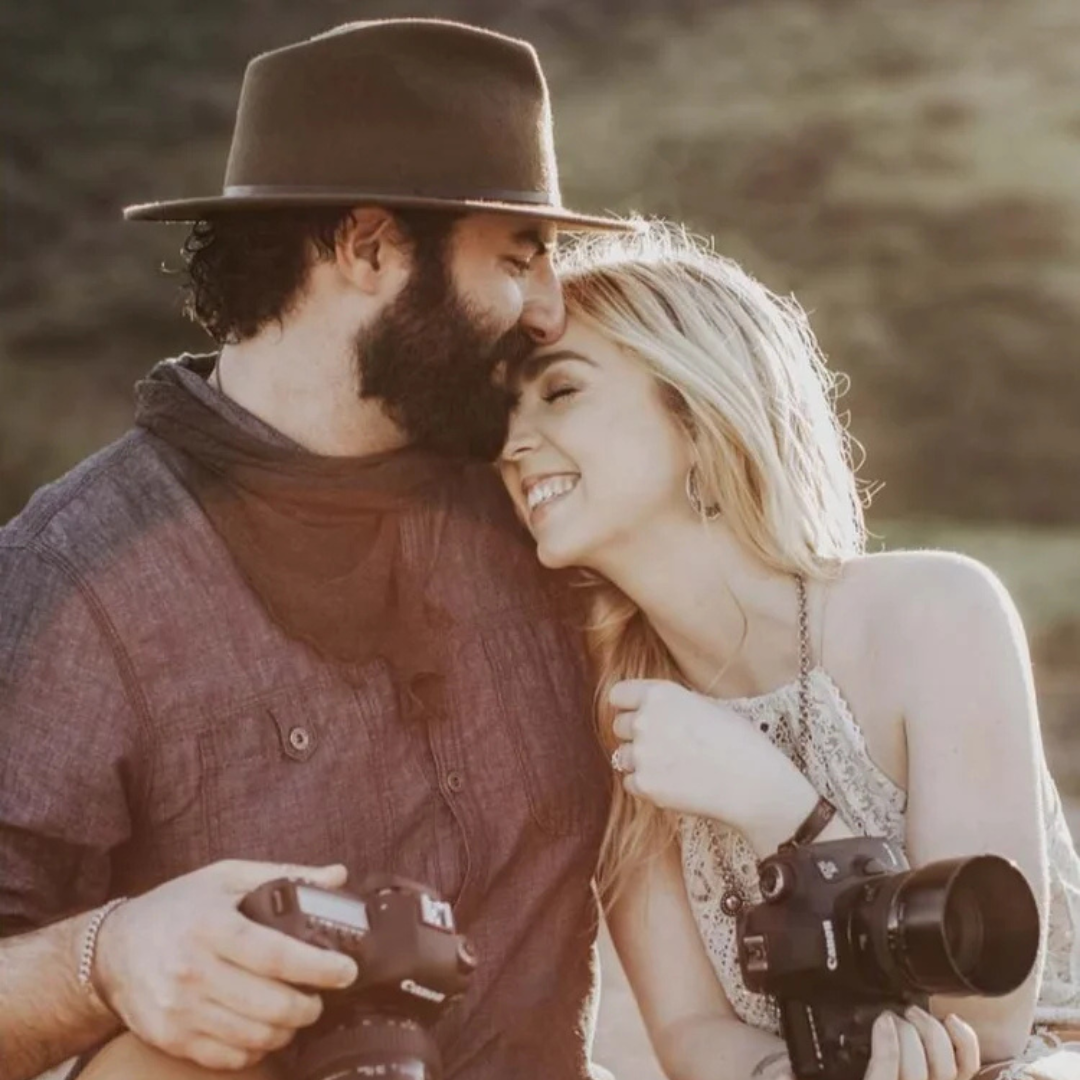 Couple smiling and touching foreheads, each holding a camera outdoors in warm sunlight